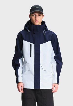 Mand iført en mørkeblå og hvid hættetrøje med lynlås foran og lommer, kombineret med sorte cargobukser og en sort kasket.