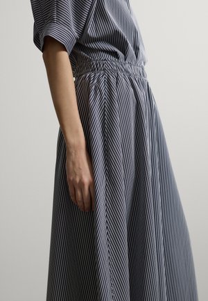 Donna che indossa un vestito a righe verticali bianche e nere, con maniche corte e vita elastica, in piedi davanti a uno sfondo chiaro e uniforme.