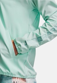 Light mint green athletic jacket featuring a smooth texture, elastic cuffs, side pocket, and embossed "UNDER ARMOUR" logo on the sleeve.