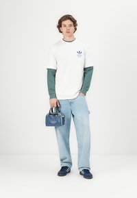 T-shirt blanc superposé sur une chemise à manches longues verte, sac à main bleu avec logo, jeans bleu clair et baskets bleues avec rayures blanches.