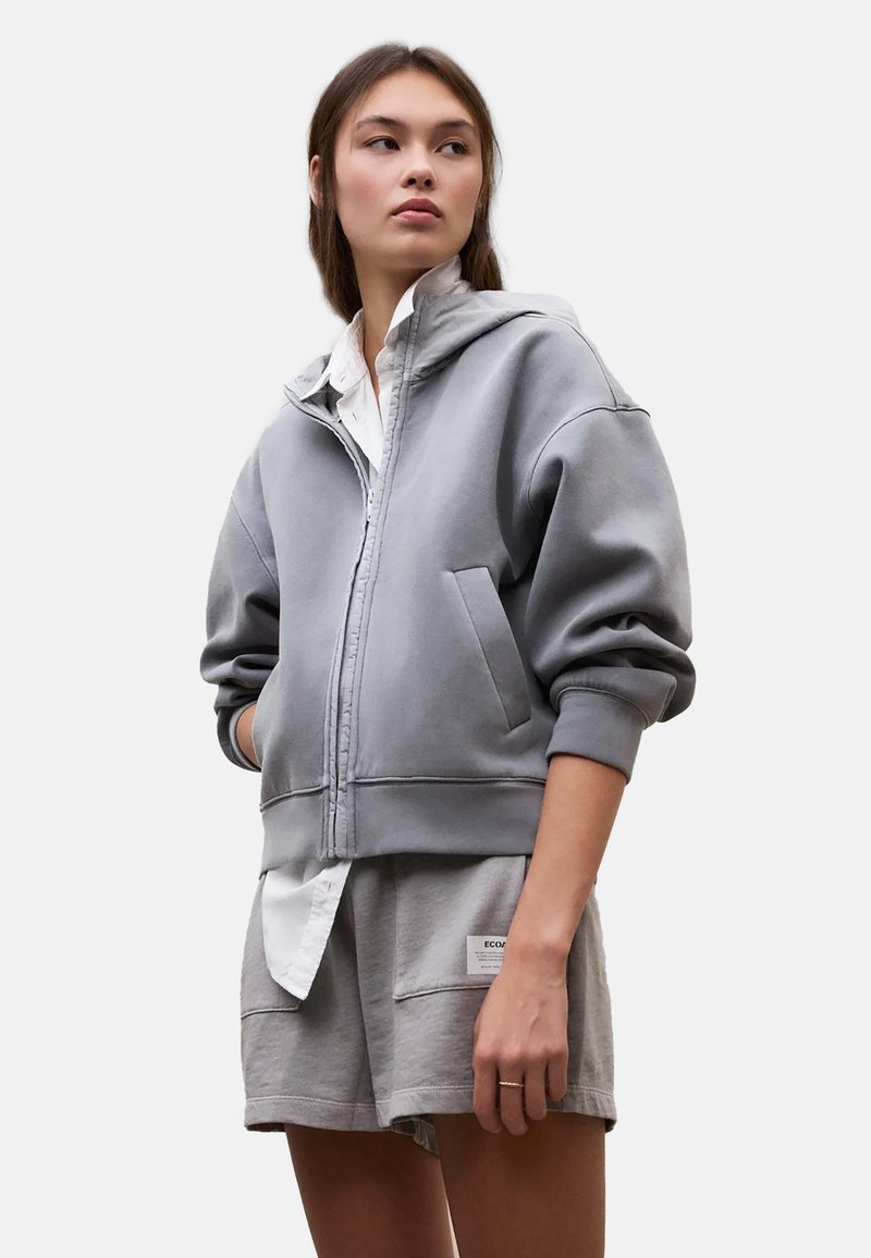 Jonge vrouw draagt een grijze hoodie met rits over een wit shirt en bijpassende grijze korte broek, staand met handen in de zakken en opzij kijkend.