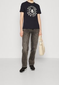 Marineblå t-shirt med hvidt logo, grå jeans med en falmet tekstur, sorte sko og en beige taske med et blødt design og håndtag.