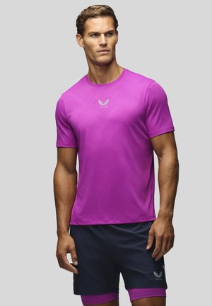 Man draagt een felpaarse atletische T-shirt met korte mouwen en zwarte short met bijpassende paarse compressieshort eronder.