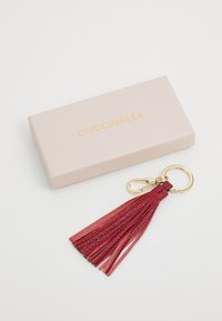 Rød lær tassel nøkkelring med gullhardware, presentert i en lyserosa boks med gull-logo. Tasselen har flere franselag.