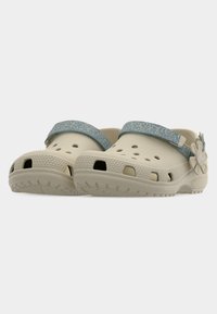 Chaussures style sabot beige avec des brides texturées bleues et un pendentif en forme de fleur, comportant des trous d'aération et des semelles épaisses en caoutchouc.