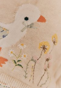 Strikket beige stof med broderet hvid fugl, orange næb, lille grøn sløjfe og broderede vilde blomster i gul, hvid og pink.