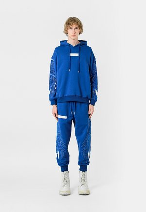 Sudadera azul con capucha y joggers a juego, con bordados en forma de plumas en las mangas y en las piernas, que cuentan con un bolsillo frontal y cordones ajustables.