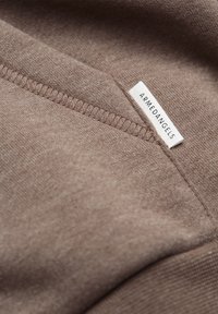 Närbild av en brun tygsöm med en vit klädkant märkt "ARMEDANGELS" sydd i kanten.