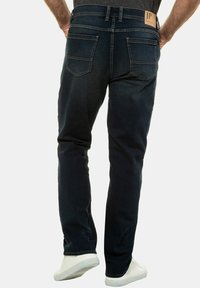 Tamsiai mėlynos džinsinės kelnes su tiesiu kirpimu, turinčios dvi galines kišenes ir rudos spalvos logotipo etikete ant juosmens. Avimos su baltais sportbačiais.