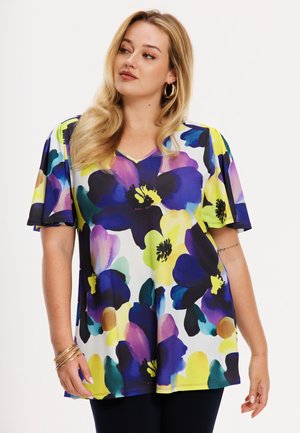 Kvinde med blondt hår iført guldringsøreringe og en kortærmet bluse med stort blomsterprint i blå, lilla, gul og grøn.