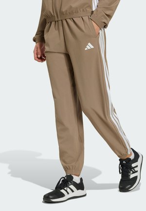 Beżowe spodnie dresowe Adidas z białymi paskami noszone przez osobę w czarno-białych butach sportowych Adidas, stojącą z jedną ręką w kieszeni.