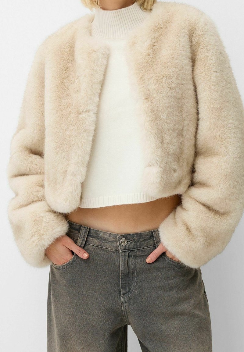 Chaqueta corta de piel sintética cremosa con cuello redondeado, diseño recortado y textura suave. Se lleva sobre un suéter de punto ligero y jeans grises.