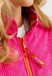 Fuksjowa kurtka z kordurowego materiału z kołnierzem stojącym, pomarańczowym zamkiem błyskawicznym oraz kolorowym wzorzystym podszewką. Posiada tkaninowy uchwyt do zaciągania.