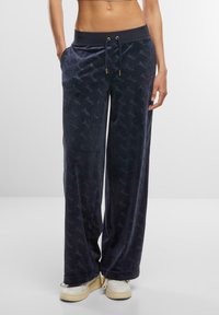 Navy velour sweatpants with a relaxed fit, featuring an elastic waistband, drawstring, and repeating logo pattern on the fabric.
