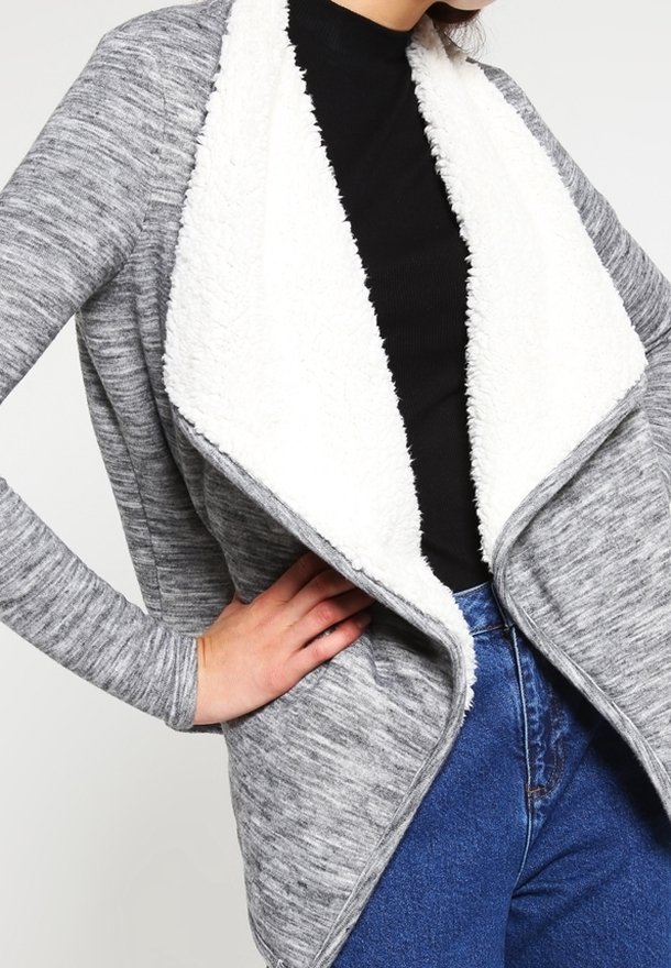 Grauer Strickcardigan mit einem weißen Fleece-Schlüsselkragen, ausgestattet mit langen Ärmeln und einem lockeren Schnitt, getragen über einem schwarzen Oberteil und blauen Jeans.