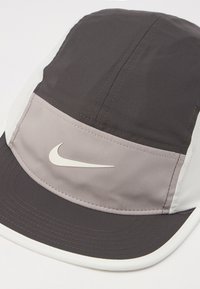Nike kapa z dvojnim odtenkom v temno sivi in svetlo sivi barvi, izdelana iz gladke tkanine s ukrivljenim robom in šivanimi detajli.