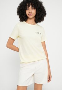 Hellgelbes Baumwoll-T-Shirt mit kurzen Ärmeln, das ein schwarzes aufgedrucktes Logo zeigt. Kombiniert mit weißen Shorts für einen lässigen Look.