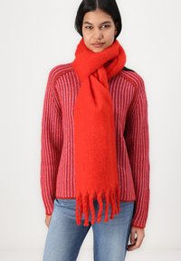 Rød ullskjerf med myk tekstur og frynsete kanter, lagd over en rød og rosa stripete strikkegenser. Enkel, uformell design.