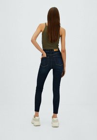 Jeans oscuros ajustados con bolsillos traseros, combinados con una camiseta sin mangas ajustada de color oliva. El modelo está de pie de espaldas, usando zapatillas de color claro.