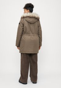 Parka castanho com capuz de pele sintética, cintura ajustável com cordão e mangas compridas. Tecido texturizado e corte reto que chega até a metade da coxa. Um par de calças de veludo cotelê castanhas é visível.