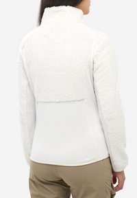 Femme portant une veste en polaire blanche avec un tissu texturé doux sur le haut du dos et les manches, associée à un pantalon beige clair, vue de dos.