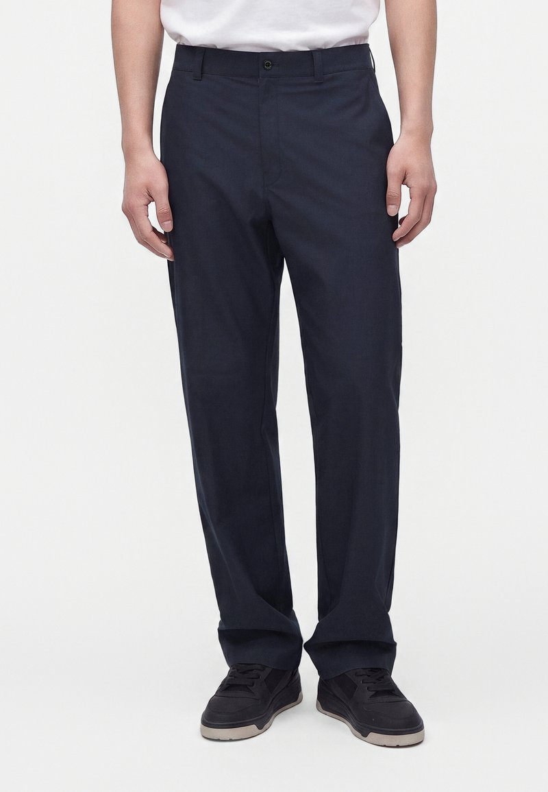 Homme portant un pantalon droit bleu marine foncé, des baskets noires et une chemise blanche, debout devant un fond clair uni.