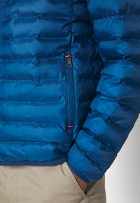 Blauwe pufferjack met horizontale stiksels, voorzien van een ritssluiting zak en een gladde, glanzende textuur. De zoom en boorden zijn elastisch.