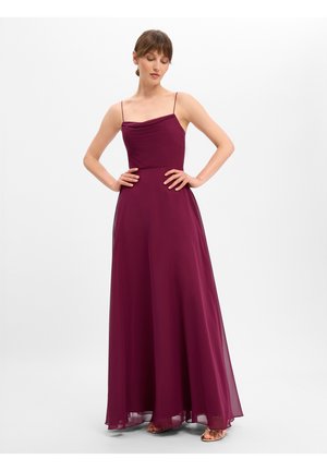 Frau mit braunen Haaren, die ein ärmelloses, bodenlanges pflaumenfarbenes Kleid mit dünnen Trägern und Sandalen trägt, steht mit den Händen in der Hüfte vor weißem Hintergrund.