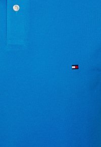 Blå polo-skjorta, bomullsmaterial, texturerad yta, två knappar vid kragen, liten broderad logotyp i röd, vit och blå på bröstet.