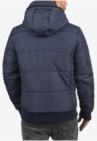 Marineblå dunjakke med hette, laget av teksturert stoff, med sømdetaljer og ribbestrikkede mansjetter. Bakbildet viser en tettsittende design.