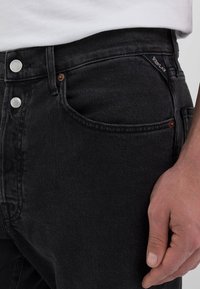 Schwarze Jeans aus Denim mit klassischem Schnitt, ausgestattet mit silberfarbenen Beschlägen und dezenten Nahtdetails. Der Stoff hat eine glatte Textur und minimale Verblassungen.