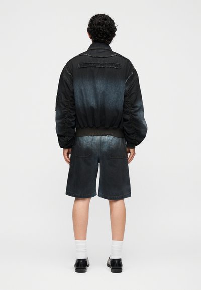 Άνδρας με σγουρά μαλλιά στέκεται με την πλάτη φορώντας μαύρο και μπλε bomber μπουφάν με διαβάθμιση χρωμάτων, σκούρα τζιν σορτς, λευκές κάλτσες και μαύρα παπούτσια.