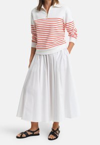 Donna che indossa una gonna bianca, una maglietta a maniche lunghe bianca con strisce orizzontali arancioni e sandali bassi neri con cinturini, in piedi con le mani nelle tasche.