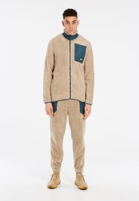 Chaqueta de forro polar beige con cierre de cremallera, cuello alto y bolsillo en el pecho color teal. Combinada con pantalones de forro polar a juego y zapatillas beige.