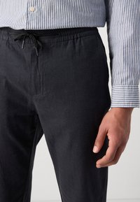 Dark navy linen trousers with an elastic waistband and drawstring, paired with a striped button-up shirt featuring a cuff.