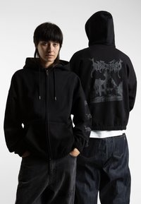 Felpa nera con zip, dotata di tasca frontale, cappuccio con coulisse e stampa grafica sul retro. Materiale in cotone, vestibilità comoda, con polsini a coste.