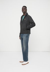 Svart jacka med dragkedja och texturerad yta, med en gul logotyp och två sidofickor, kombinerad med ljusblå jeans och vita sneakers.