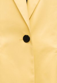Blazer jaune avec un unique bouton noir à quatre trous, fixé au centre devant sous les revers.