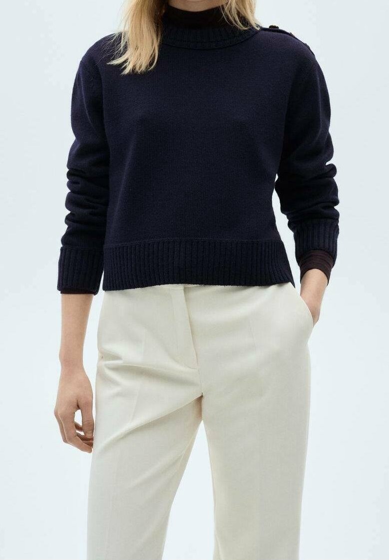 Personne portant un pull à col roulé bleu marine avec des poignets et un ourlet côtelés, associé à un pantalon taille haute de couleur crème, une main dans la poche.