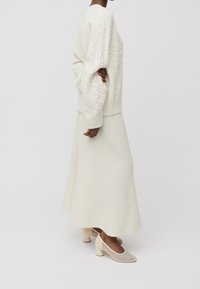 Suéter blanco de punto trenzado con mangas abullonadas, combinado con una falda larga blanca y zapatos de tacón cuadrado en color beige. Textura suave y diseño minimalista.