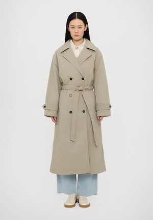 Khaki trench coat with a belted waist, large lapels, double-breasted buttons, and rolled sleeves, paired with light blue wide-leg jeans and beige shoes.