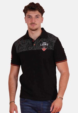 Jeune homme aux cheveux courts et bouclés portant un polo noir avec un empiècement à motifs et des détails rouges, debout devant un fond uni.