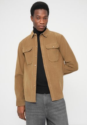 Bruine corduroy blouse met lange mouwen, knoopsluiting aan de voorkant en twee borstzakken. Gedragen over een zwarte coltrui, gecombineerd met grijze jeans.