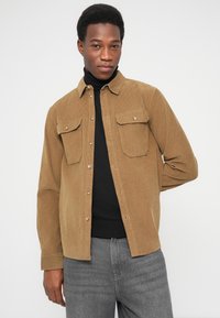 Chemise en velours côtelé marron à manches longues, boutonnée à l'avant, avec deux poches poitrine. Portée sur un col roulé noir, associée à un jean gris.