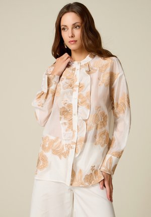 Frau trägt eine weiße Bluse mit beiger Blumenstickerei und Rüschendetail, kombiniert mit weißen Hosen, und posiert vor neutralem Hintergrund.