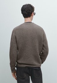 Maglione marrone a coste con maniche lunghe, collo tondo e tessuto strutturato, indossato sopra una camicia scura. Vista da dietro.