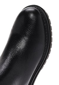 Bota de cuero negro con una textura suave, punta redondeada y una suela de goma de bajo perfil que presenta un patrón acanalado. Detalles cosidos a lo largo del borde.
