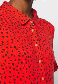 Červená košile s krátkým rukávem a černým skvrnitým vzorem, límečkem a oranžovými knoflíky. Tkanina vypadá měkce a má jemnou strukturu.