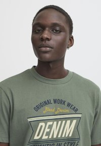 Zelené bavlněné tričko s kulatým výstřihem, s modrým a žlutým grafickým textovým designem "DENIM" a "Original Work Wear." Měkká textura.