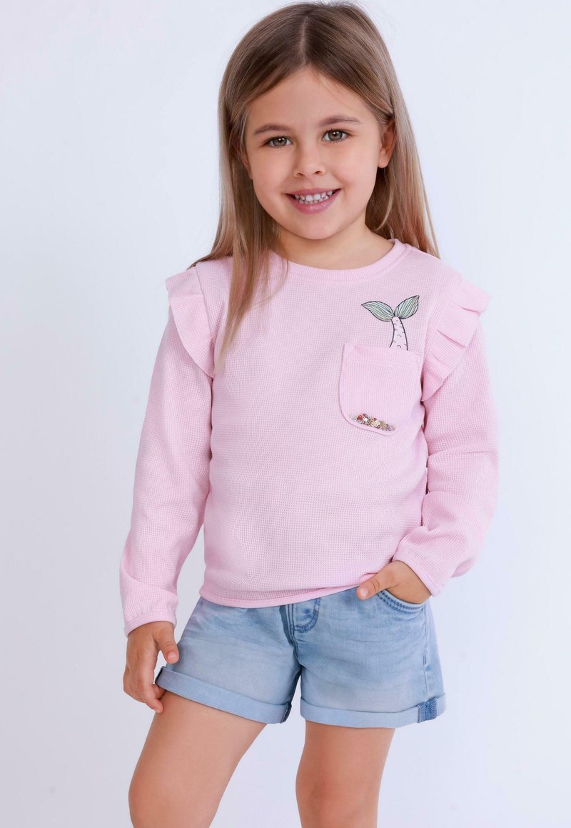 Ljusrosa långärmad topp med textilstrukturer, volangdetalj vid axlarna, ficka med en broderad växt, kombinerad med jeansshorts.